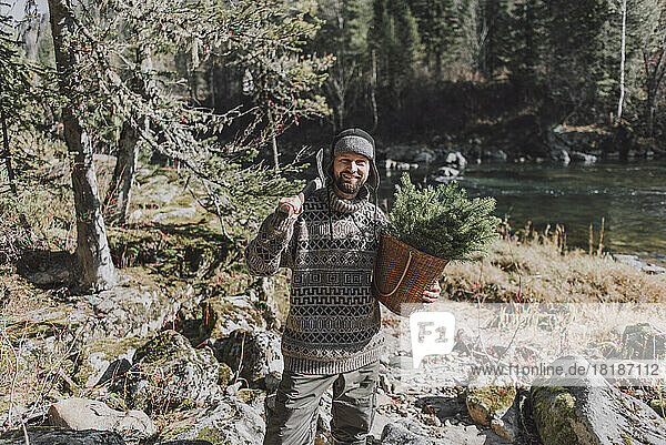 Man holding axe and basket with twigs of spruce tree on sunny day