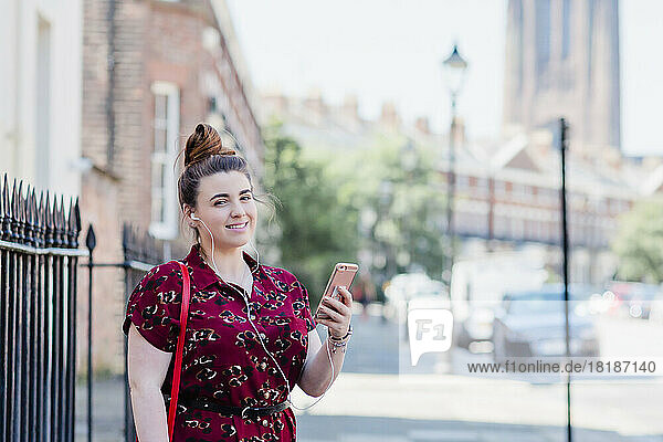 Portrait of woman standing on pavement using earphones and cell phone  Liverpool  UK
