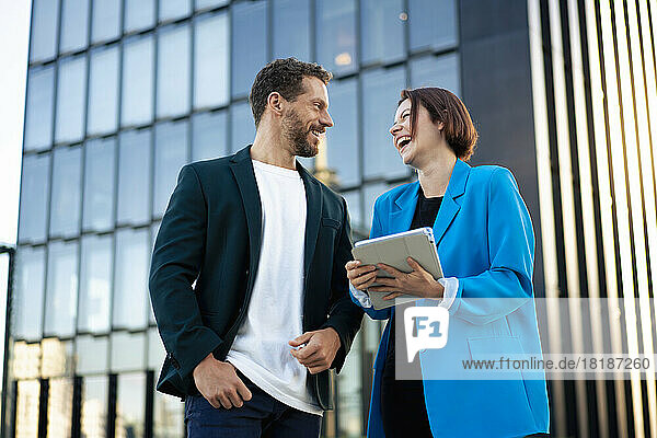 Happy businesswoman holding tablet PC talking with colleague in front of building