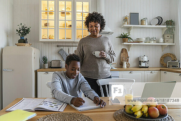 Junge Frau mit Kaffeetasse schaut ihrem Sohn dabei zu  wie er Notizen auf dem Tisch in der Küche schreibt