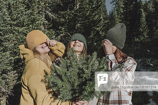 Happy friends covering eyes with knit hats in forest