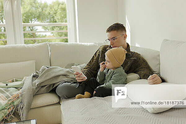 Young man using smart phone with son sitting on sofa at home