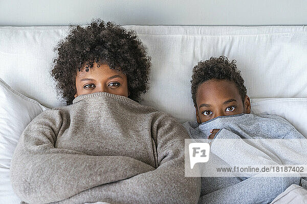 Mother and son covering mouth with sweater lying on bed at home