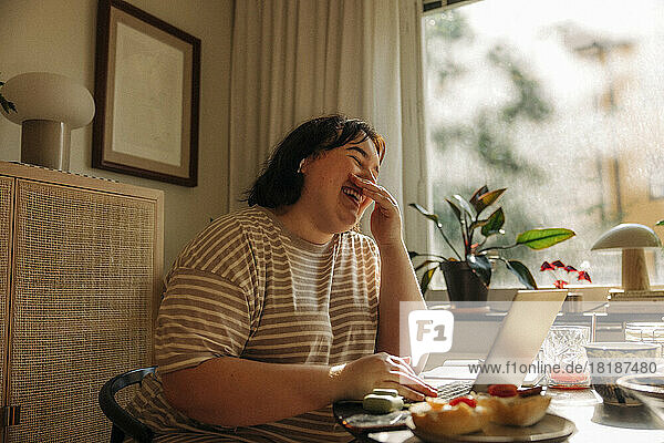 Fröhliche junge Frau  die den Mund mit den Händen bedeckt  während sie am Tisch am Fenster sitzt