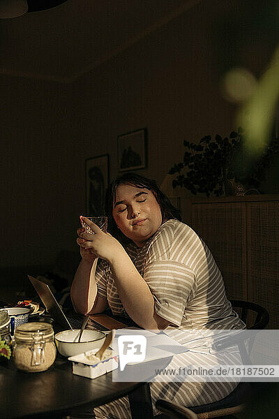 Junge Frau mit geschlossenen Augen  die ein Trinkglas hält  während sie am Tisch sitzt