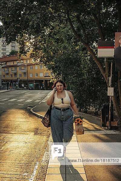 Frau mit Einkaufstasche in voller Länge  die bei Sonnenuntergang auf dem Fußweg mit ihrem Smartphone telefoniert