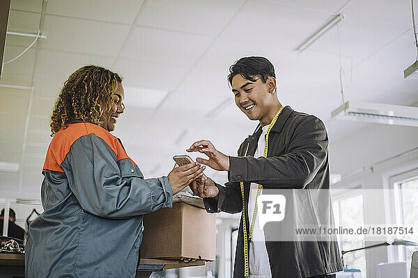 Lächelnder männlicher Modedesigner  der eine digitale Unterschrift leistet  während er ein Paket von einem Zusteller in einer Werkstatt entgegennimmt