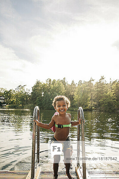 Smiling shirtless boy holding railing while standing on jetty near lake during vacation