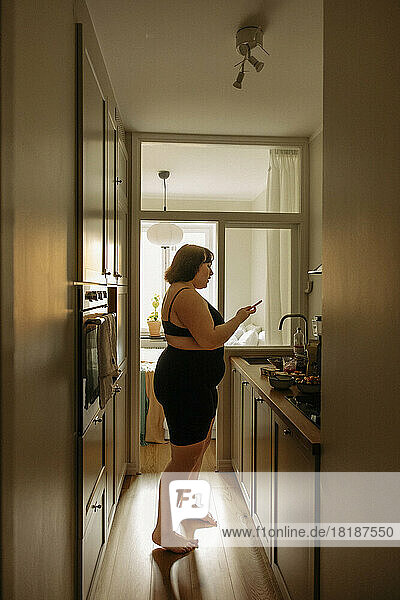 Seitenansicht einer jungen  üppigen Frau  die ihr Smartphone in der Küche zu Hause benutzt