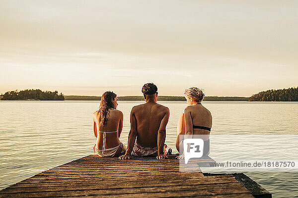 Rückansicht von männlichen und weiblichen Freunden  die im Urlaub auf einem Steg am See sitzen