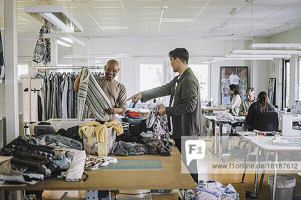 Seitenansicht eines männlichen Modedesigners  der auf einen Kollegen zeigt  der in einer Werkstatt einen Pullover hält