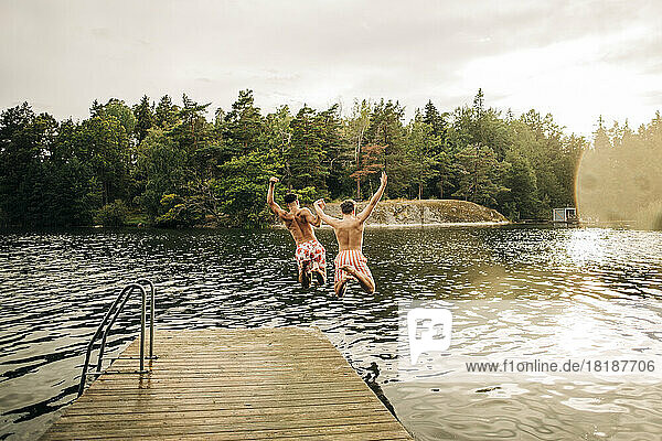 Male friends with arms raised jumping in lake from jetty during vacation