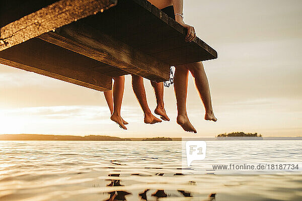 Low section of male and female friends sitting on jetty by lake during sunset