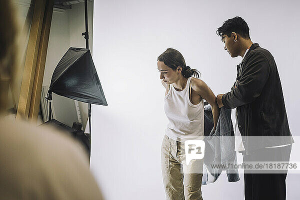 Männlicher Designer hilft einem Modemodell beim Tragen einer Jacke während eines Fotoshootings im Studio