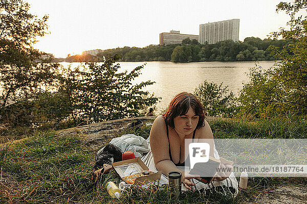Young voluptuous woman reading book while lying against lake