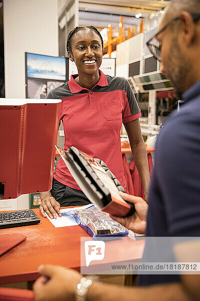 Lächelnde weibliche Kassiererin  die einen männlichen Kunden betrachtet  der eine Ware an der Kasse hält