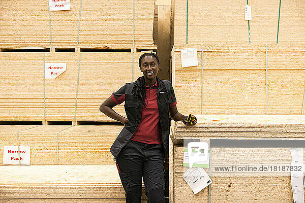 Portrait of smiling saleswoman with hand on hip standing near plywood stack at hardware store
