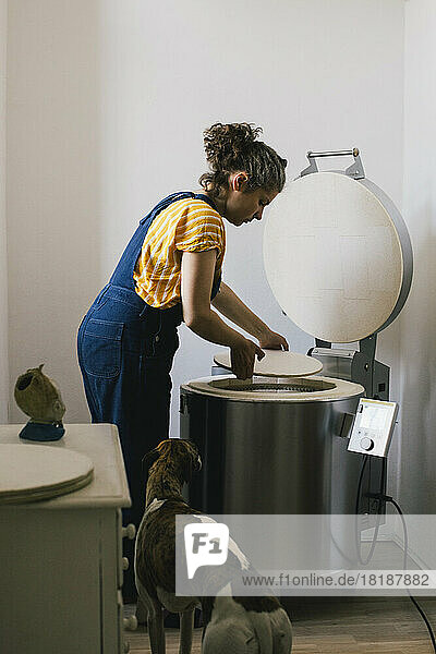 Seitenansicht einer Handwerkerin  die bei ihrer Arbeit zu Hause eine Keramikplatte in einen elektrischen Brennofen legt