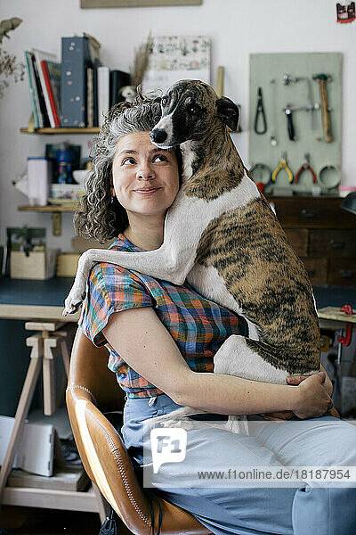 Lächelnde Frau  die ihren Hund umarmt  während sie zu Hause auf einem Stuhl sitzt