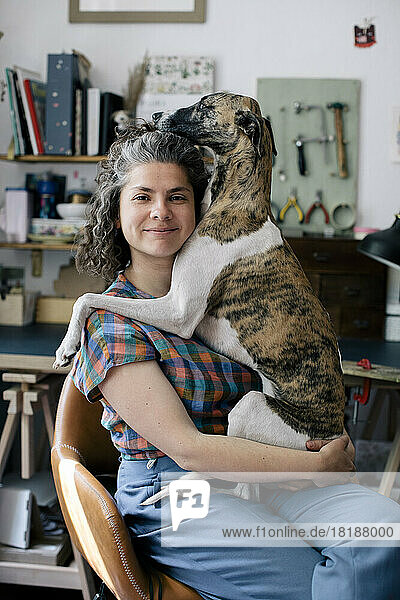 Porträt einer lächelnden Frau  die ihren Hund umarmt  während sie zu Hause auf einem Stuhl sitzt
