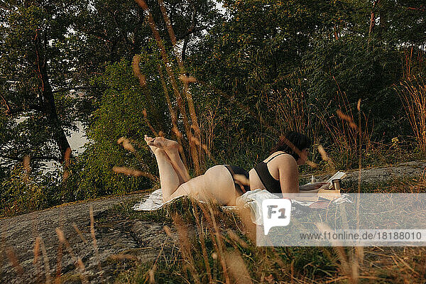 Junge Frau im Bikini in voller Länge  die ein Buch liest  während sie an einem Baum liegt
