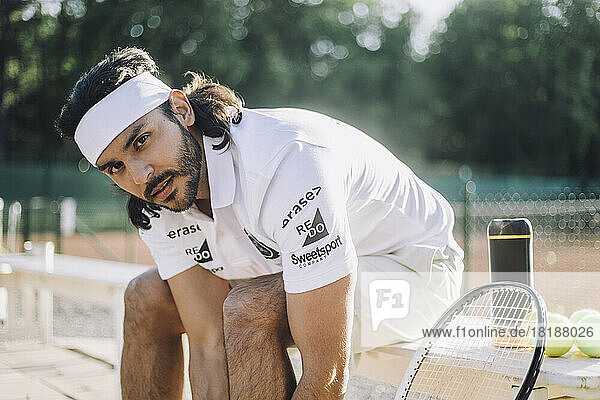 Selbstbewusster Mann mit Stirnband  der auf einer Bank auf dem Tennisplatz sitzt
