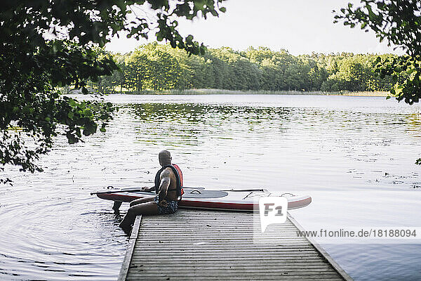 Mann mit Paddelboard schaut auf den See  während er auf dem Steg sitzt