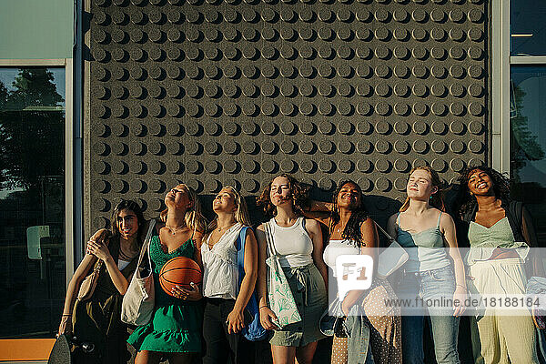 Smiling teenage girls with eyes closed enjoying sunny day while standing against wall