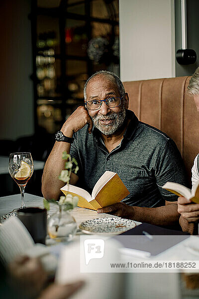 Älterer Mann mit Brille sitzt mit Buch in einem Cafe