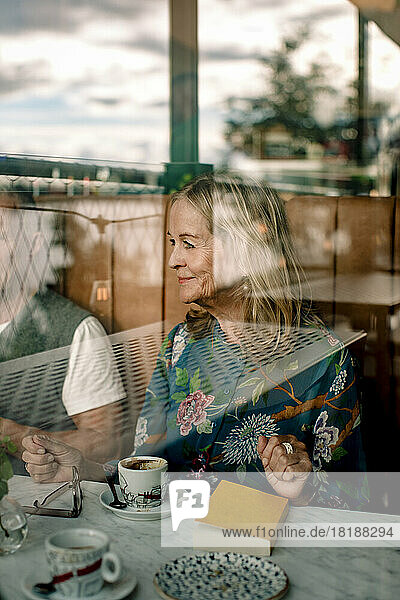 Lächelnde ältere Frau  die mit einem Buch bei einem männlichen Freund in einem Café sitzt  gesehen durch Glas