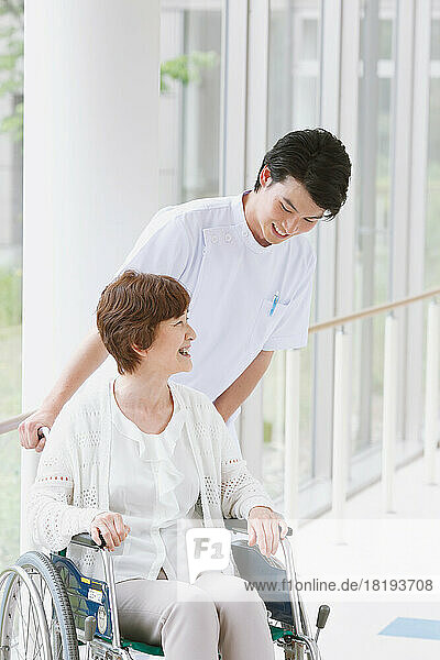 A senior Japanese woman in a wheelchair and a young male nurse having a conversation in the hallway