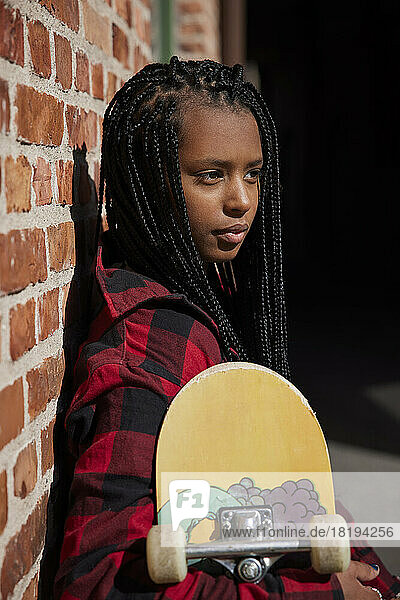 Teenage girl holding skateboard while leaning on brick wall