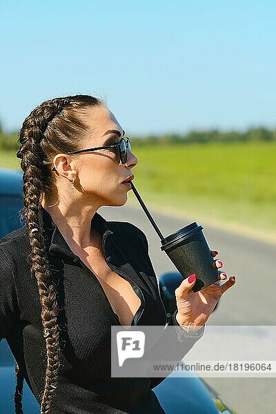 Frau trinkt starken Kaffee durch den Strohhalm aus einem Becher zum Mitnehmen in der Nähe ihres Autos