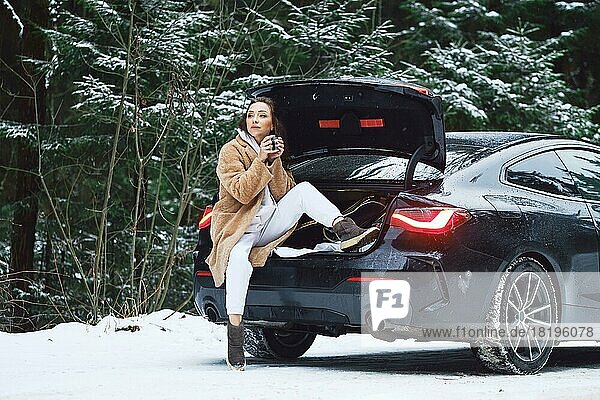 Eine müde Autofahrerin hält ihr Auto auf einem winterlichen Waldweg an  um sich auszuruhen und eine Tasse Kaffee zu trinken