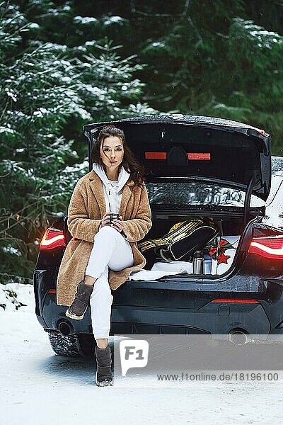 Woman sits on a car trunk with gift box inside and drinks hot coffee in winter forest road