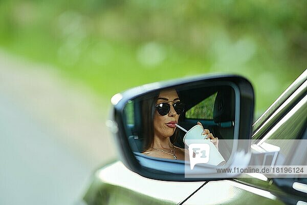Spiegelung einer Frau  die ein Getränk im Rückspiegel eines Autos trinkt
