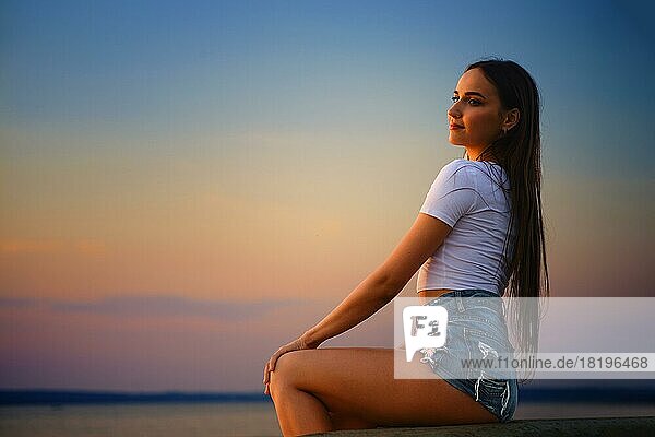 Junge Frau sitzt im Profil auf einem Pier und beobachtet den Sonnenuntergang