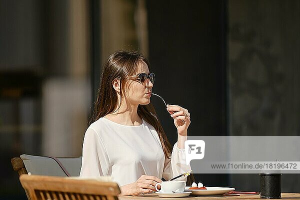 Schöne junge Frau mit Sonnenbrille sitzt auf der Terrasse des Cafés in sonnigen Tag und essen Kuchen