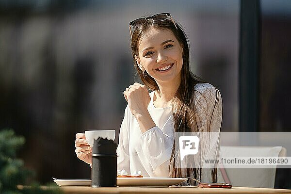 Hübsche junge Frau mit einer Tasse Cappuccino in der Hand schaut geradeaus und lächelt