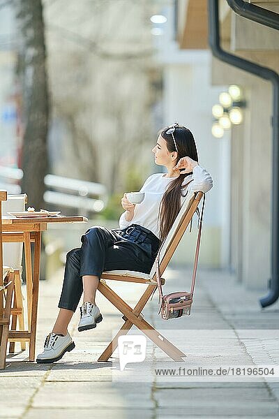 Pensive young woman sitting on the terrace of the cafe in sunny day and drinking coffee