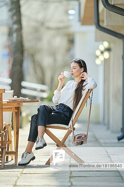 Pensive young woman sitting on the terrace of the cafe in sunny day and drinking coffee