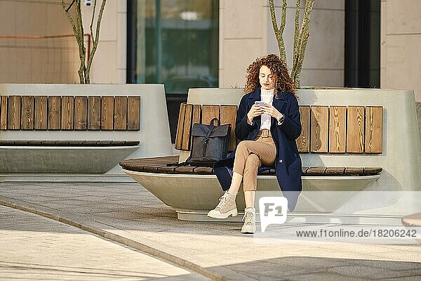 Ernste Frau  die auf einer Bank sitzt und Nachrichten auf ihrem Smartphone im Freien überprüft