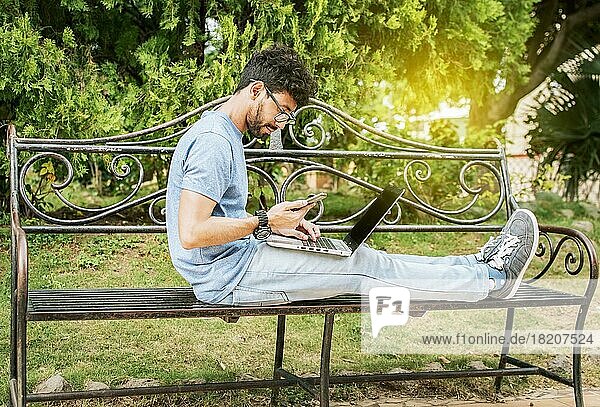 Freelancer man sitting in a park using laptop and cellphone. Man in a park working online with laptop. Relaxed man working with laptop outdoor  Young man in a park with laptop and cell phone