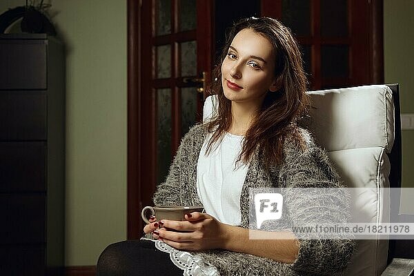 Happy smiling girl with big cup of tea in her hands on armchair in room