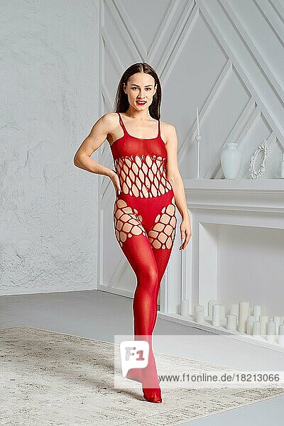 Wunderschöne junge Frau im roten Ganzkörper-Fischnetz-Bodysuit posiert im Studio mit einer Hand auf der Taille