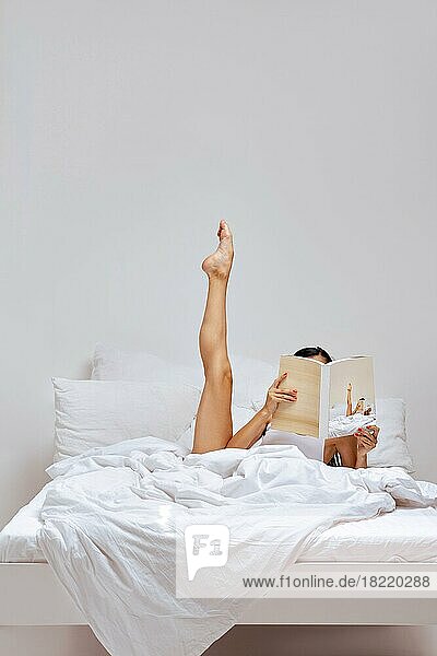 Junge Frau im Bett versteckt Gesicht mit einem Buch und streckt ihr Bein aus