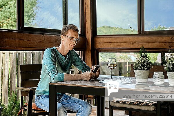 Ein lächelnder Mann mittleren Alters trinkt Wein  während er auf der Veranda seines Landhauses sitzt und sein Smartphone benutzt