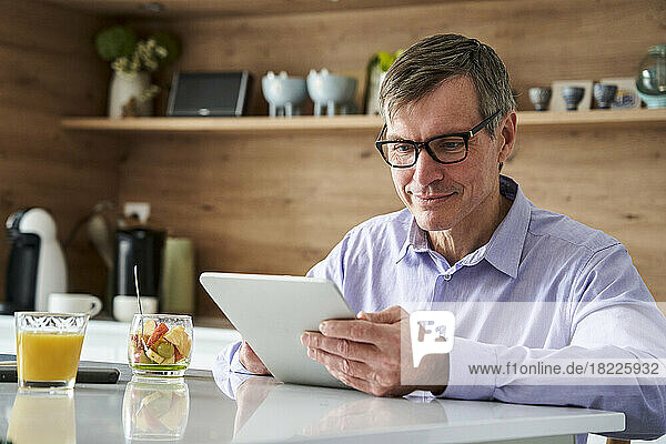 Middle-aged professional man smiling and checking messages on digital tablet while having breakfast on kitchen counter at home