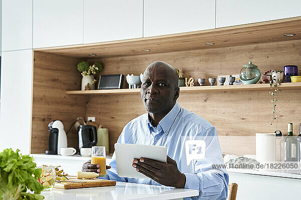 Senior African-American man sitting in kitchen eating fruit for breakfast while checking messages on digital tablet