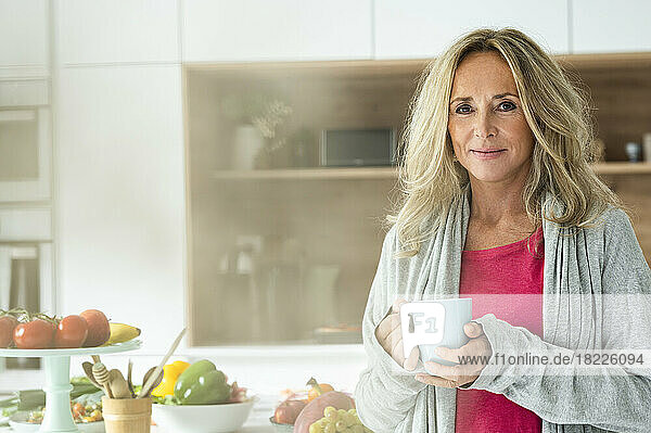 Portrait of middle-aged woman holding a cup of coffee looking at camera standing in kitchen